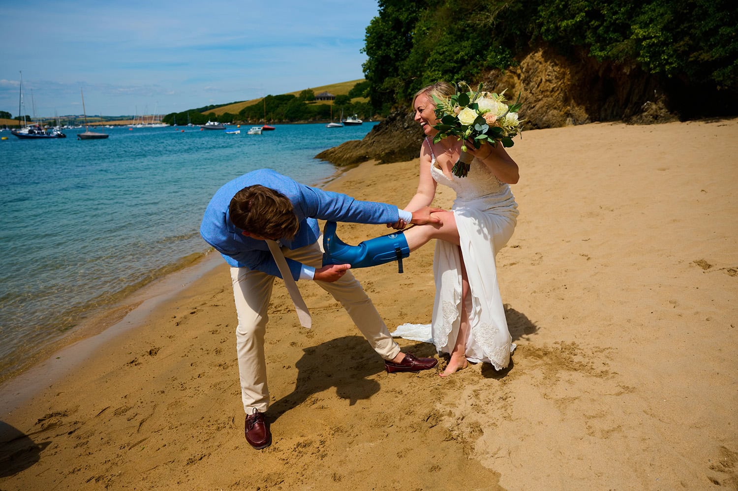 bride and grrom on beach in salcombe with groom pulling wellie boot off the bride in full laughter on sunny summer day