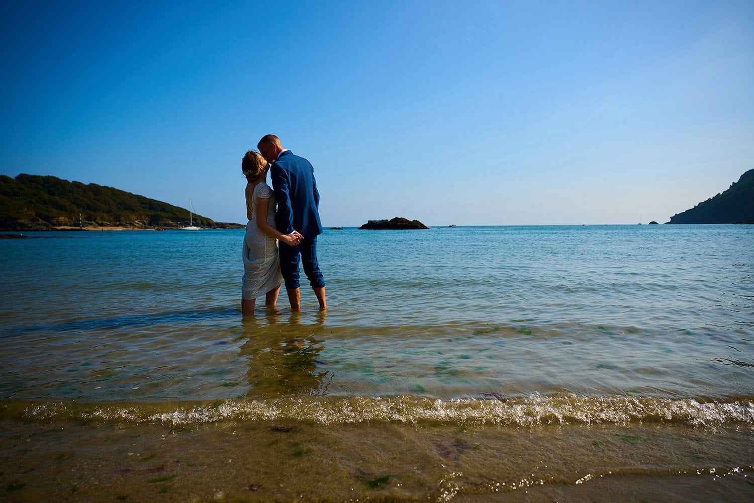 bride and groom standing in the sea with shoes off kissing with blue skies during summer wedding