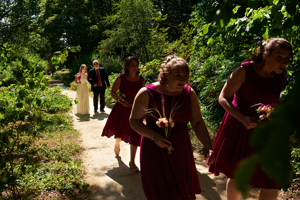 here comes the bride- walking through trees to outside wedding in dappled light