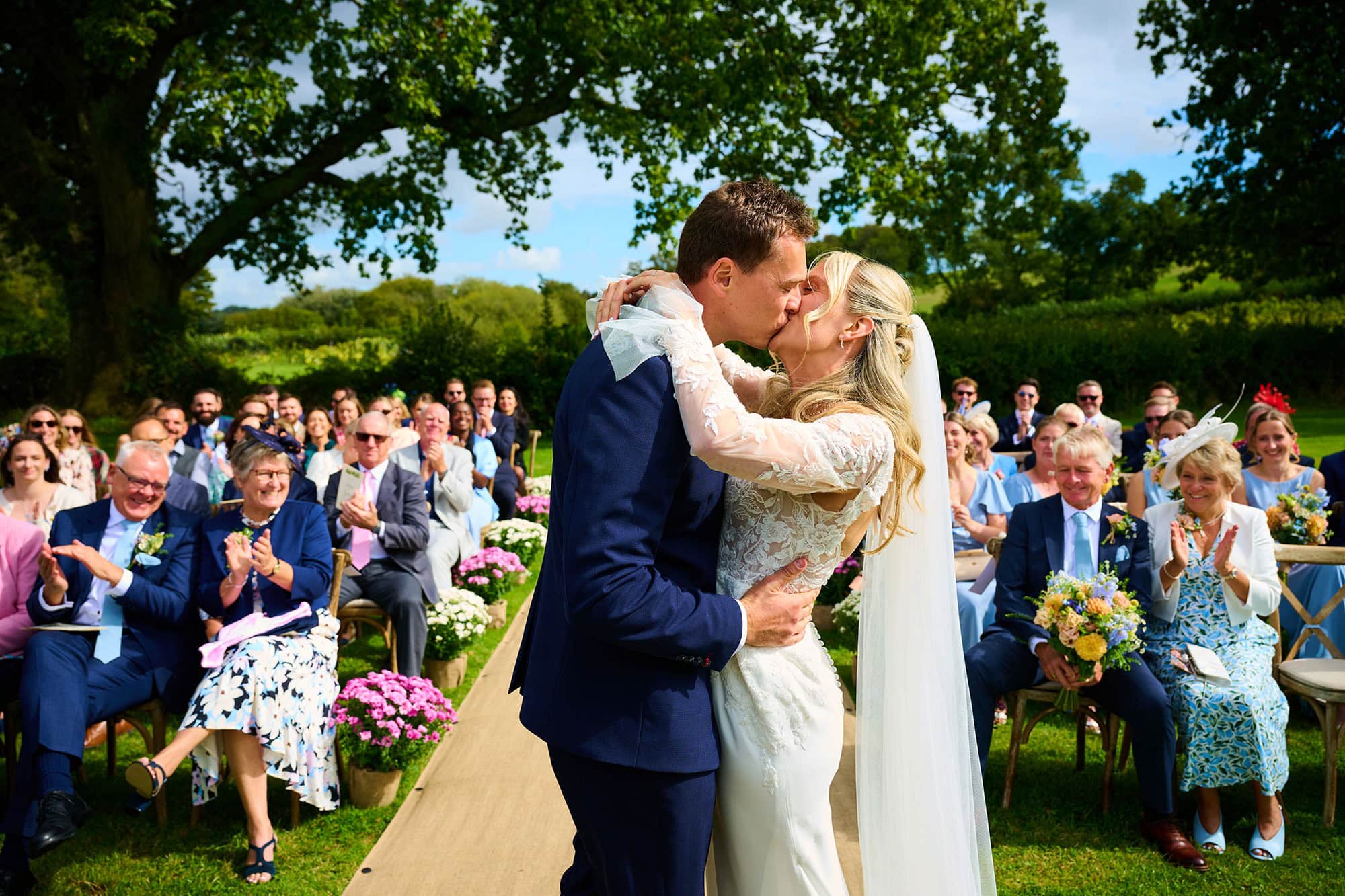 bride and groom's first kiss during their outside ceremony at English Oak Vineyard with blue skies and guests looking on