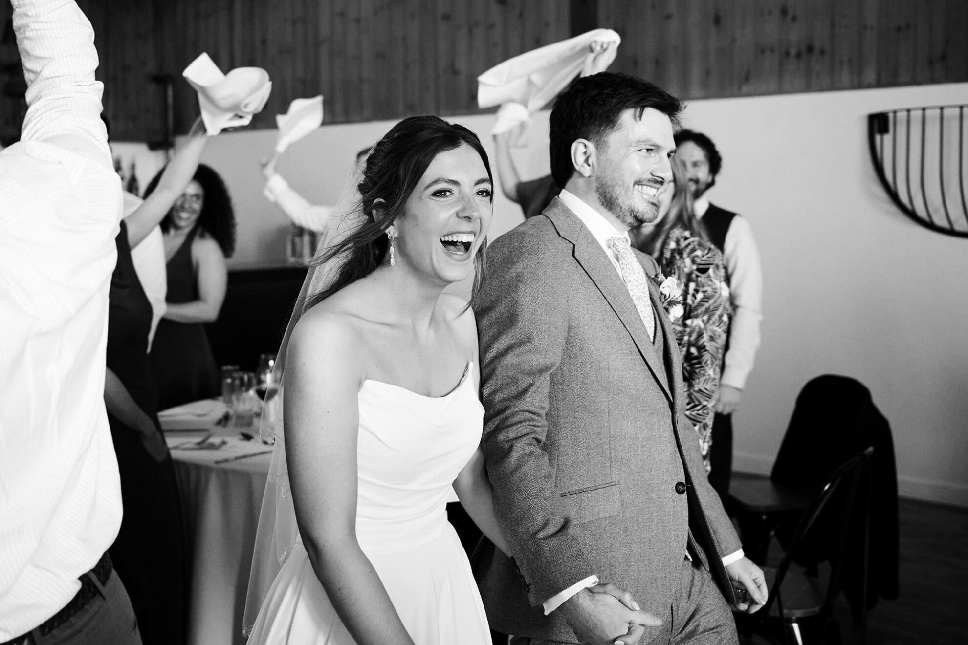 Upton Barn wedding in Devon with bride and groom entering wedding breakfast laughing with waving napkins in black and white